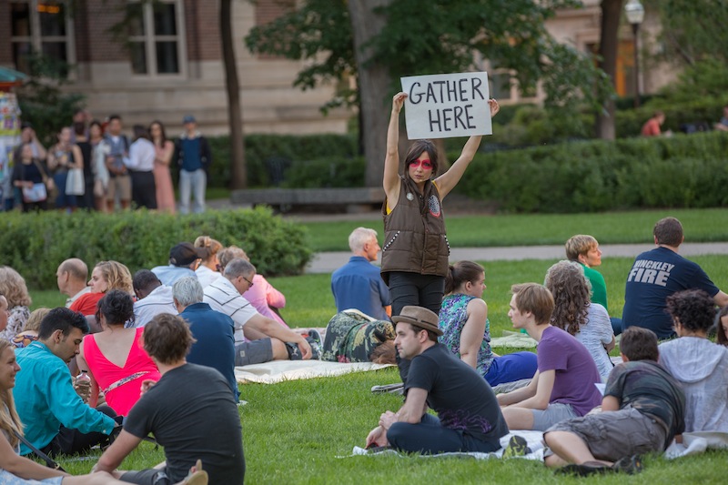 Emily Johnson holding a sign that says: gather here amongst a group of people sitting on grass